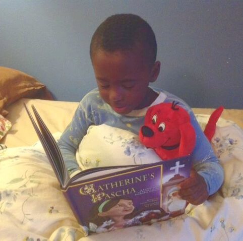 A child sitting in bed and reading Catherine's Pascha, with a plush red dog under his arm "reading" with him.