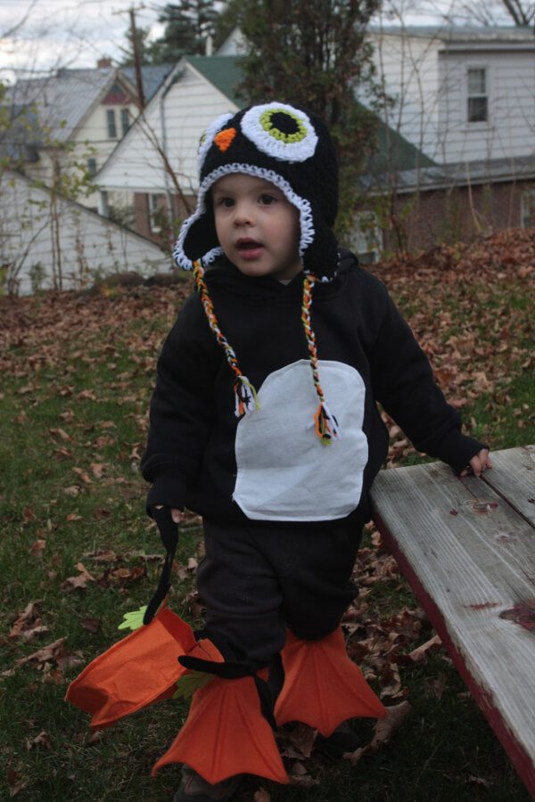 A small cild in a Halloween costume standing in his back yard. The costume is a penguin, made from a black sweatsuit and swimming flippers.