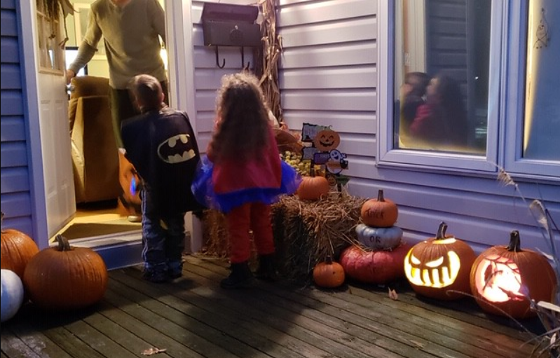 a boy in a batman costume and a girl in a Wonder Woman costume at an open door, where and adult offers candy